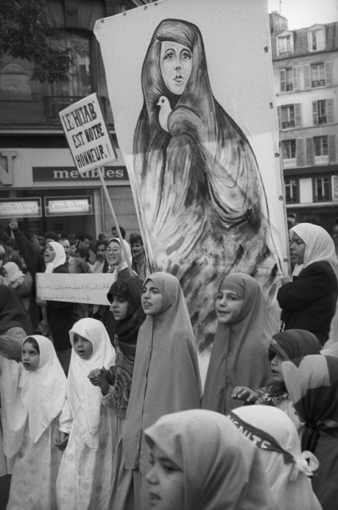 1989年10月22日、クレイユの中学校での「ヴェール事件」を受け、パリで公立学校のスカーフ着用規制に抗議するイスラム教徒のデモ。約400人の参加者の中で、「ヒジャブはあなたの名誉」と掲げる女性たちが先頭に立つ。デモへの参加・不参加を含め、ムスリムコミュニティ内でも立場は一枚岩ではなかった。 写真:A. Abbas/Magnum Photos/アフロ